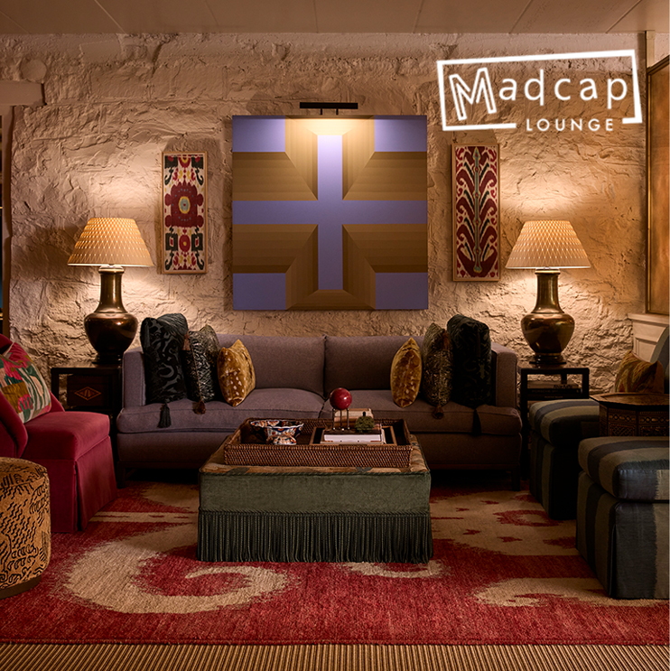 Cozy lounge with eclectic decor, patterned lamps, plush couches, bold rug, central ottoman, and abstract art on a stone wall. Madcap Lounge sign in the top right corner.