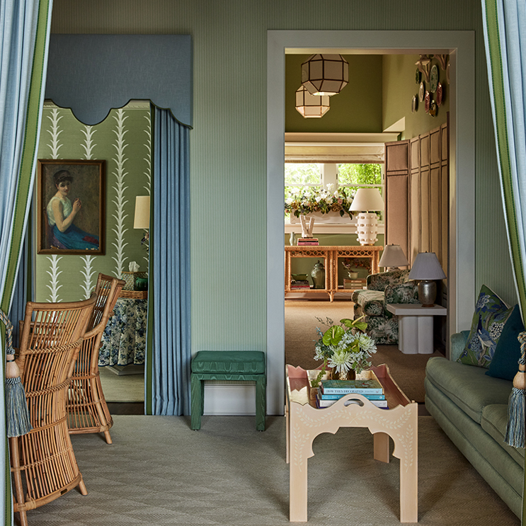 A cozy, green-toned living space with wicker chairs, a sofa, a scalloped mirror, floral wallpaper, and layered rooms decorated with lamps, plants, and geometric light fixtures.