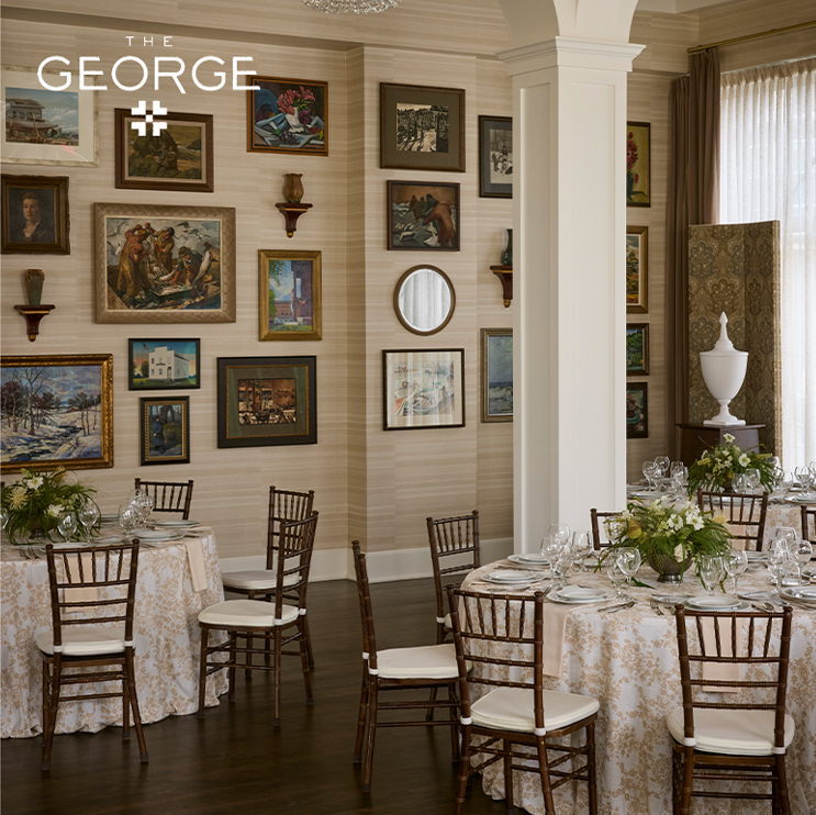 Elegant dining room at The George, featuring round tables with white floral tablecloths, wooden chairs, and walls adorned with framed art and mirrors. Large windows with sheer curtains let in natural light.