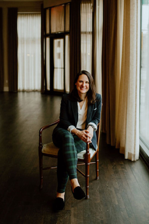 A smiling woman wearing a dark blazer, white top, and plaid trousers sitting in a wooden chair.
