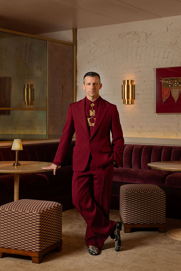 A man wearing a burgundy suit and patterned shoes stands in a restaurant with velvet booths.